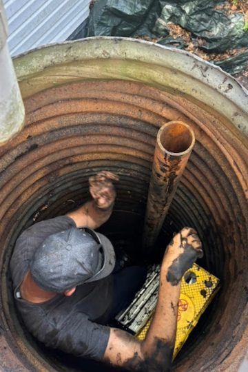 The image shows a person wearing a gray cap and a gray shirt working inside a large, circular, corrugated metal pit or well. The person is covered in mud, especially on their arms, suggesting they are performing a dirty maintenance or repair task. There is a rusty vertical pipe in the middle of the pit and a yellow crate with holes sitting on the ground beside the person. The walls of the pit are rust-colored with dark stains, and some pipes and green tarps are visible outside the pit above ground. The environment suggests a plumbing, septic, or drainage repair scenario.
