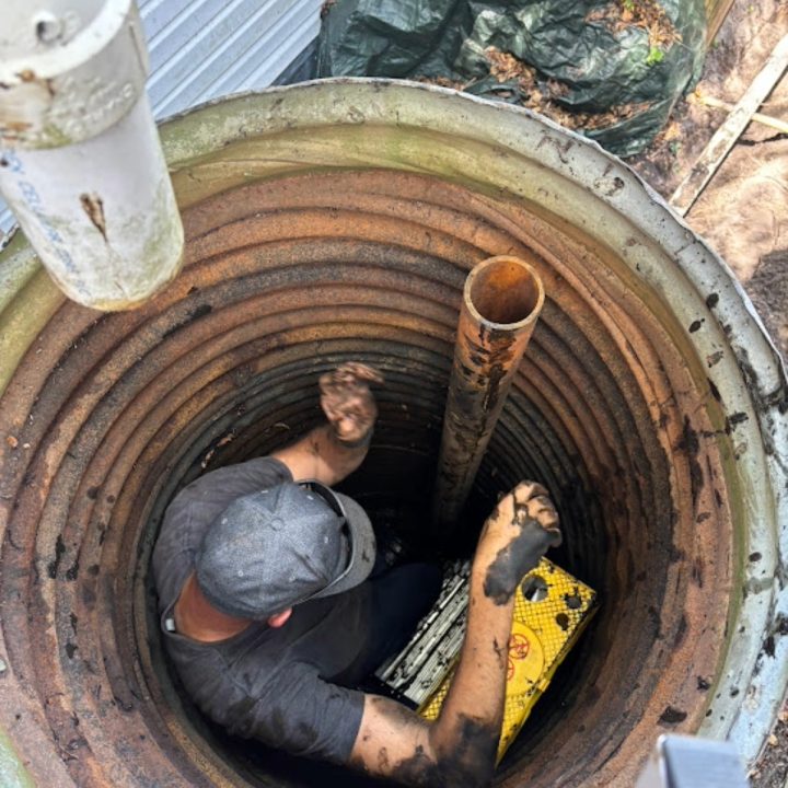 The image shows a person wearing a gray cap and a gray shirt working inside a large, circular, corrugated metal pit or well. The person is covered in mud, especially on their arms, suggesting they are performing a dirty maintenance or repair task. There is a rusty vertical pipe in the middle of the pit and a yellow crate with holes sitting on the ground beside the person. The walls of the pit are rust-colored with dark stains, and some pipes and green tarps are visible outside the pit above ground. The environment suggests a plumbing, septic, or drainage repair scenario.