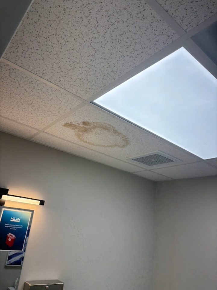 Water-stained ceiling tile in commercial restroom with light panel and vent nearby, showing need for ceiling repair