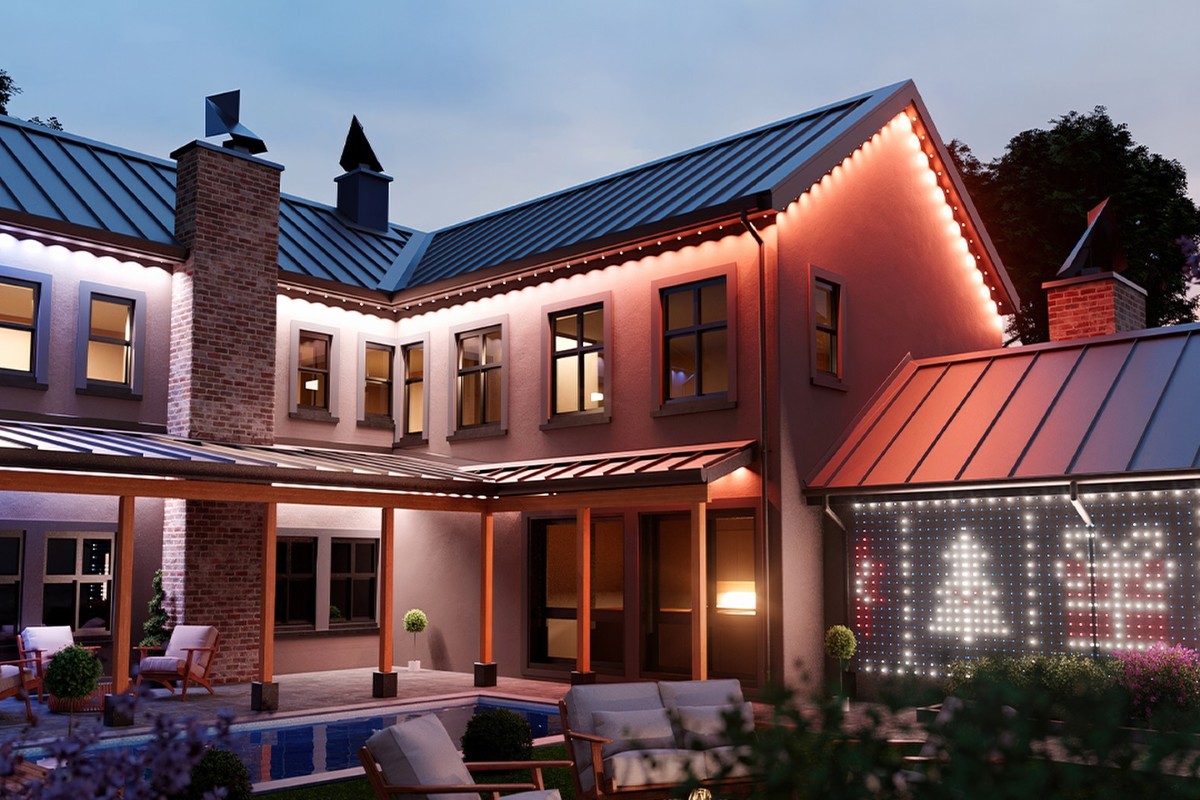 A modern two-story home with a sleek metal roof and brick chimney, beautifully decorated with warm white Christmas lights outlining the roofline. The patio area features cozy outdoor seating around a small pool, and a nearby wall displays festive LED patterns of a Christmas tree and gift box, creating a stylish and inviting holiday atmosphere at dusk.