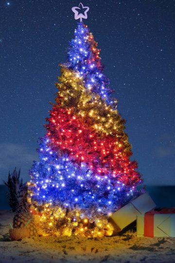 A beautifully lit Christmas tree on a sandy beach at night, decorated with layers of golden, red, and blue lights and topped with a glowing star. Presents sit at the base of the tree next to a pineapple with straws, while a starry sky and the dark ocean provide a serene tropical backdrop.