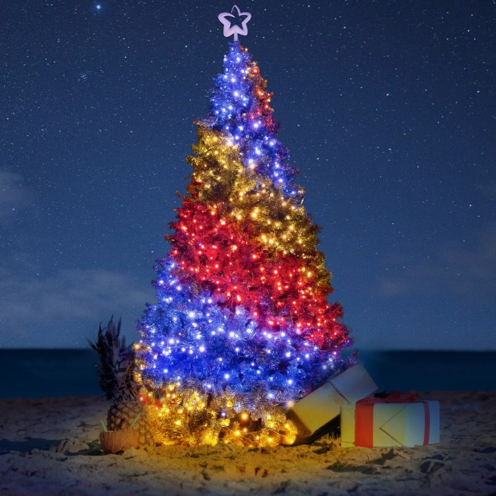 A beautifully lit Christmas tree on a sandy beach at night, decorated with layers of golden, red, and blue lights and topped with a glowing star. Presents sit at the base of the tree next to a pineapple with straws, while a starry sky and the dark ocean provide a serene tropical backdrop.