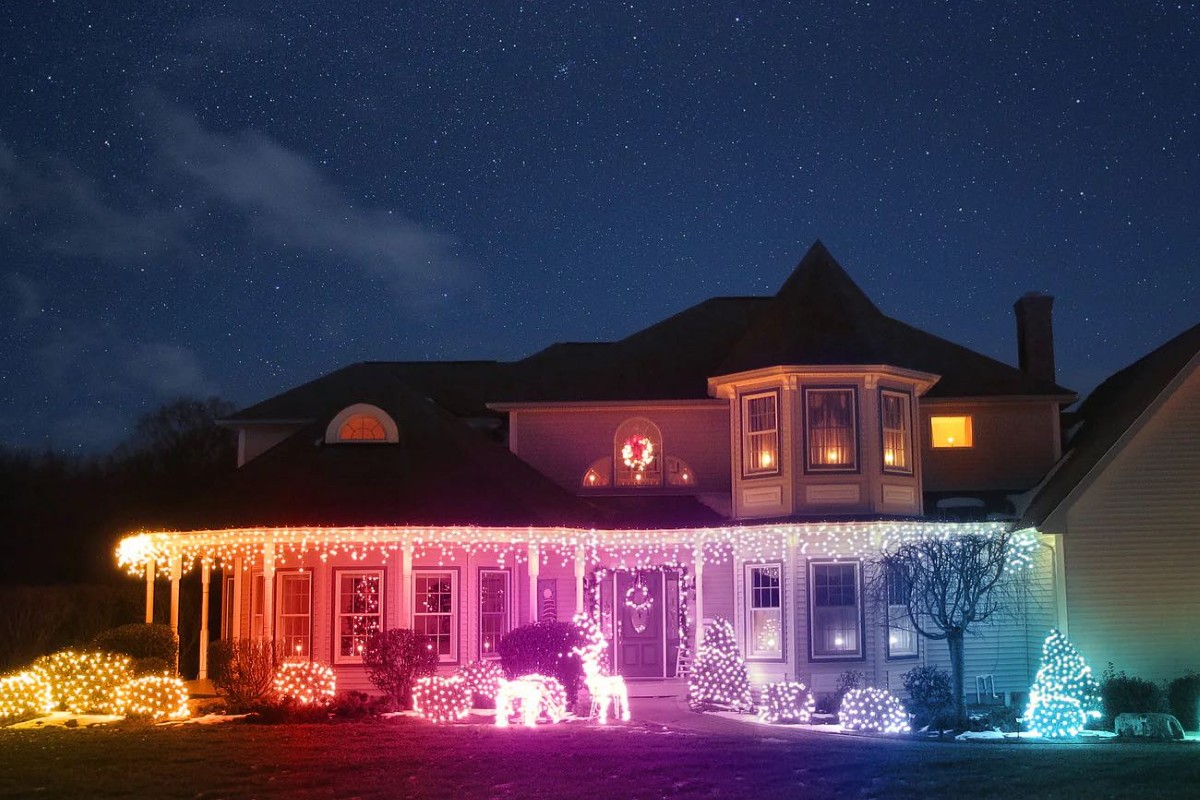 A large two-story house beautifully decorated for Christmas, with cascading icicle lights along the roofline and porch. The yard features glowing reindeer and bushes wrapped in colorful lights that shift from warm yellow and pink to cool blue and white. A lit wreath hangs above the front door, and the house glows softly under a clear, star-filled night sky.